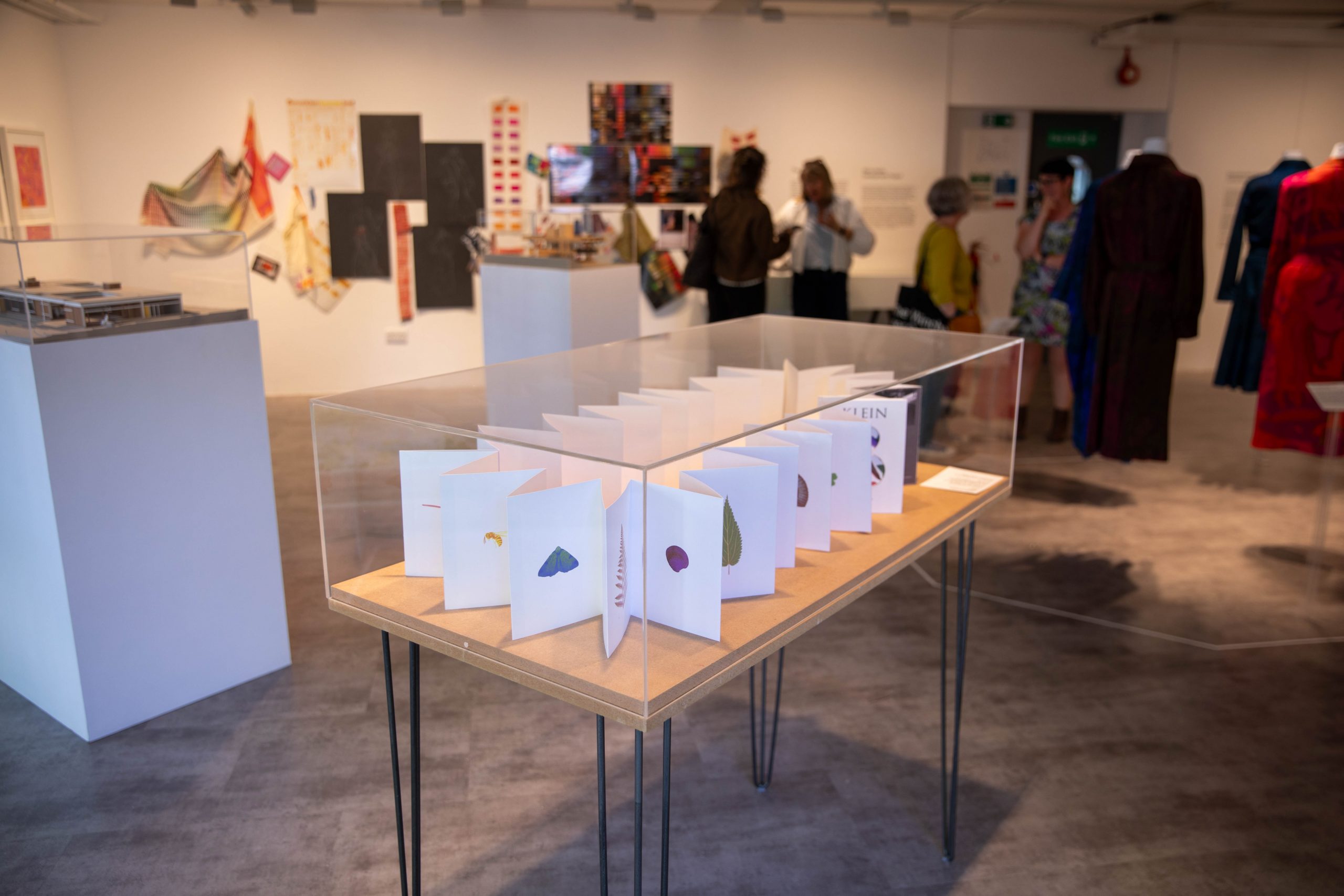 A display case containing an artist’s book or folded publication is shown within a gallery exhibition. Fashion pieces, textile works and visitors are visible in the background.