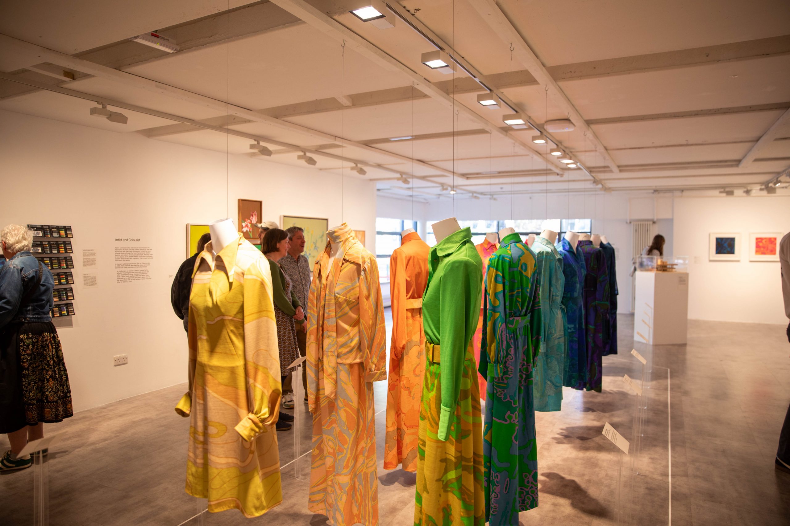 A gallery exhibition displays brightly coloured patterned garments suspended on headless mannequins arranged in a row. Visitors view the clothing and artworks throughout the exhibition space.