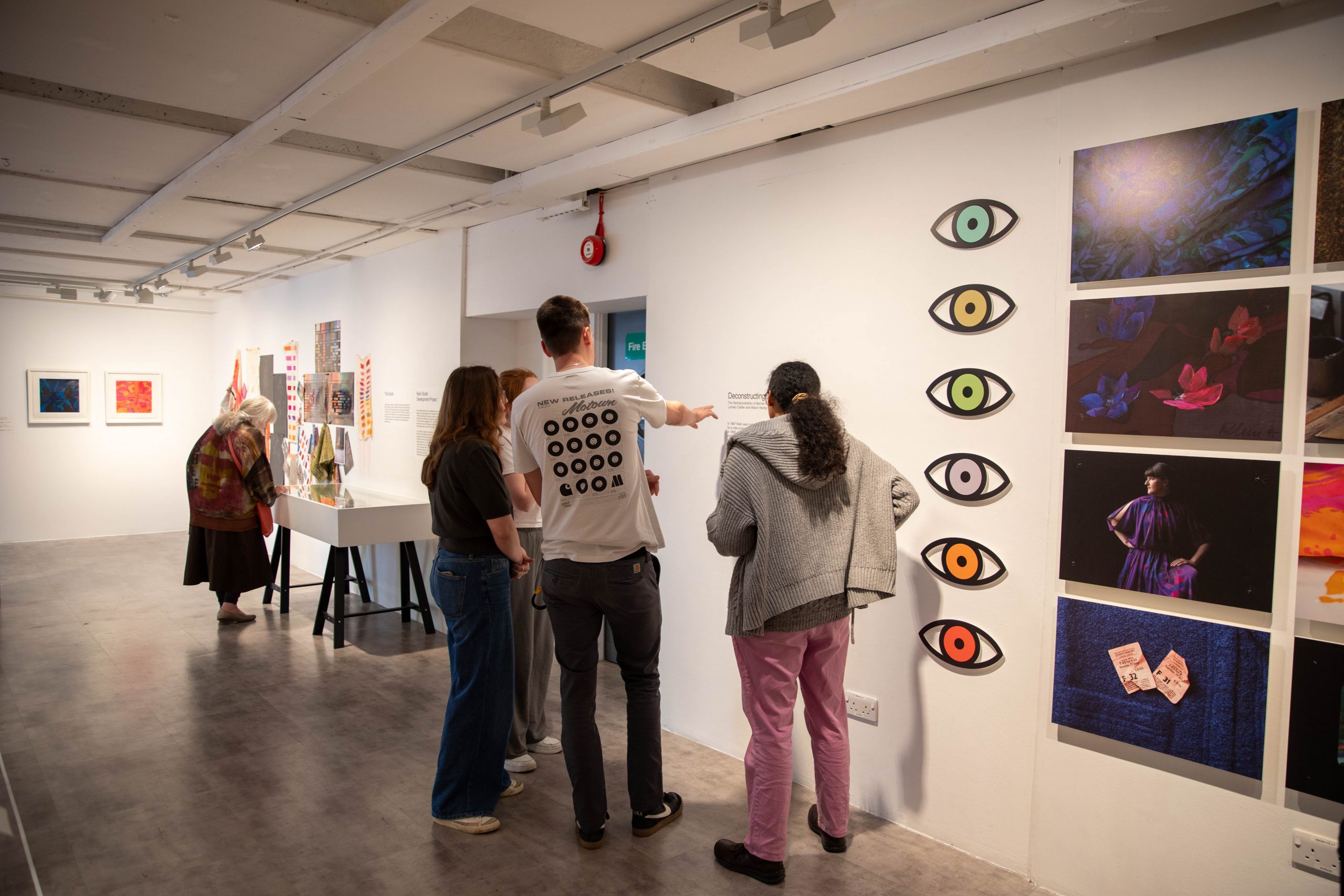 Visitors view a gallery wall display featuring photographs, graphic artworks and eye-shaped motifs arranged in a vertical sequence. Additional exhibition displays and interpretation panels are visible throughout the space.