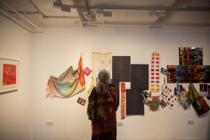 A visitor examines a mixed-media display of textile samples, colour studies, sketches and design materials mounted on a gallery wall. The exhibition presents elements of the creative and design development process.