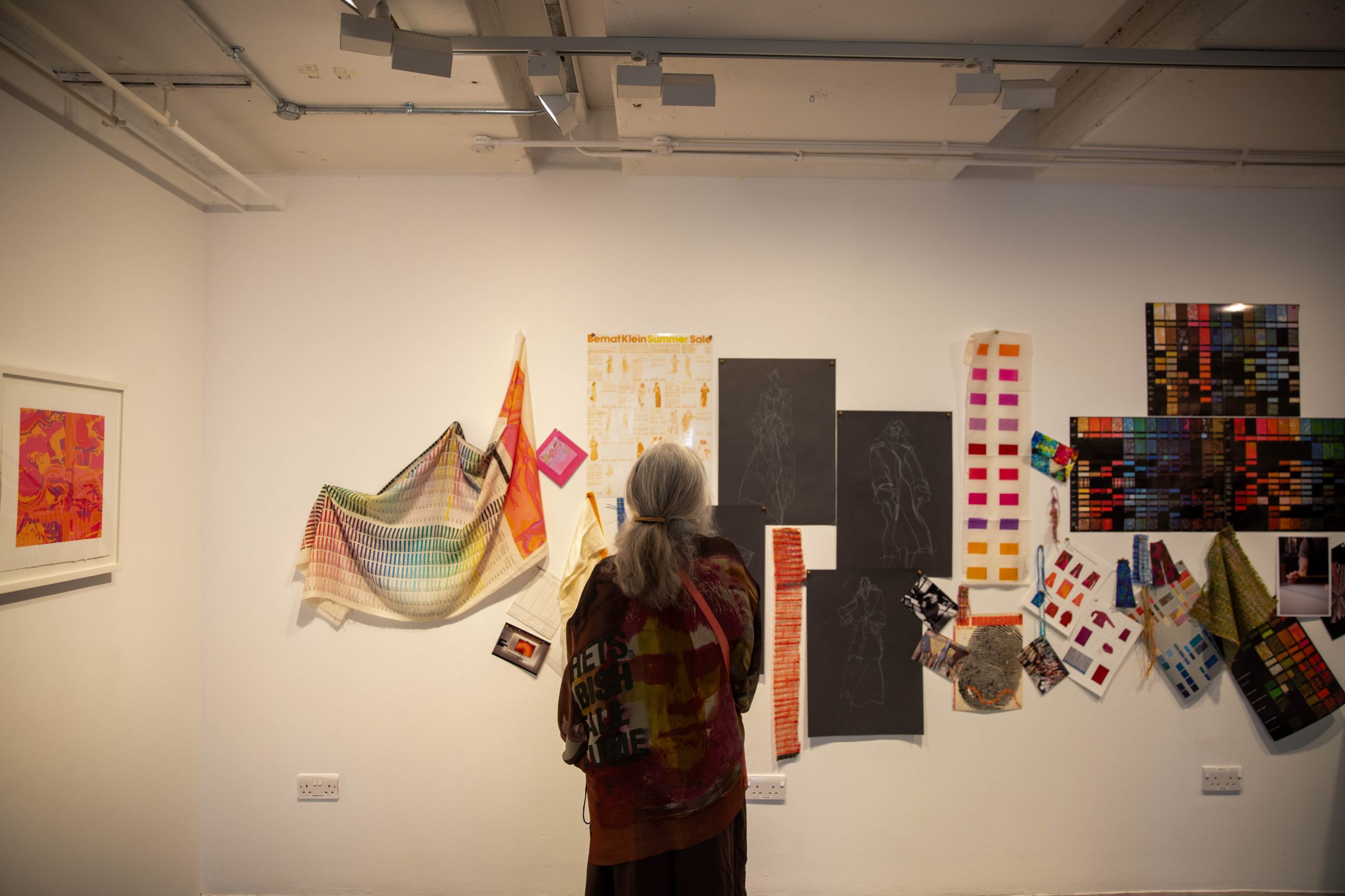 A visitor examines a mixed-media display of textile samples, colour studies, sketches and design materials mounted on a gallery wall. The exhibition presents elements of the creative and design development process.