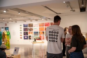 Visitors converse within a gallery exhibition featuring suspended garments, photographs and display cases. Large photographic prints and colourful fashion pieces are visible in the background.