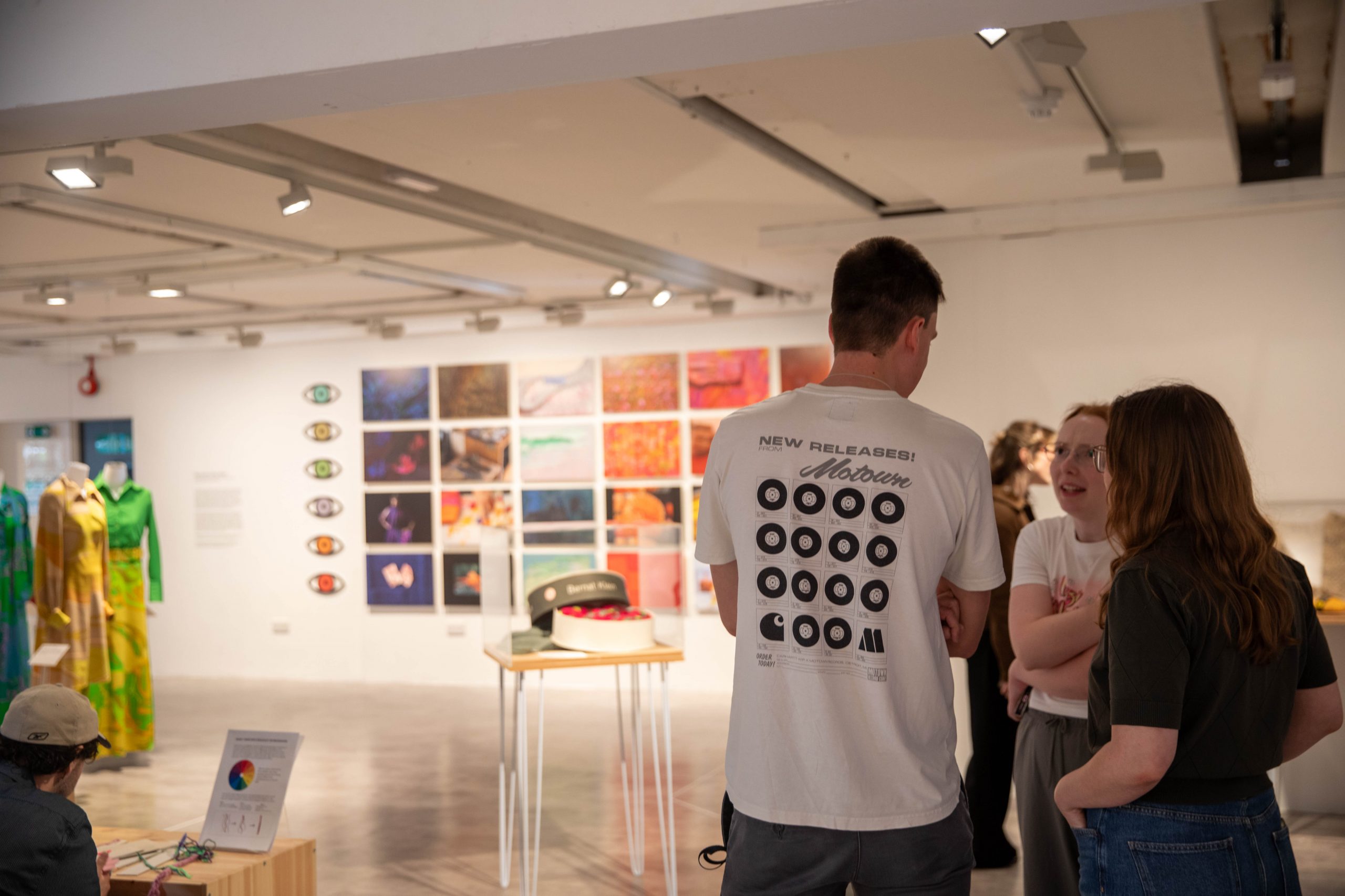 Visitors converse within a gallery exhibition featuring suspended garments, photographs and display cases. Large photographic prints and colourful fashion pieces are visible in the background.