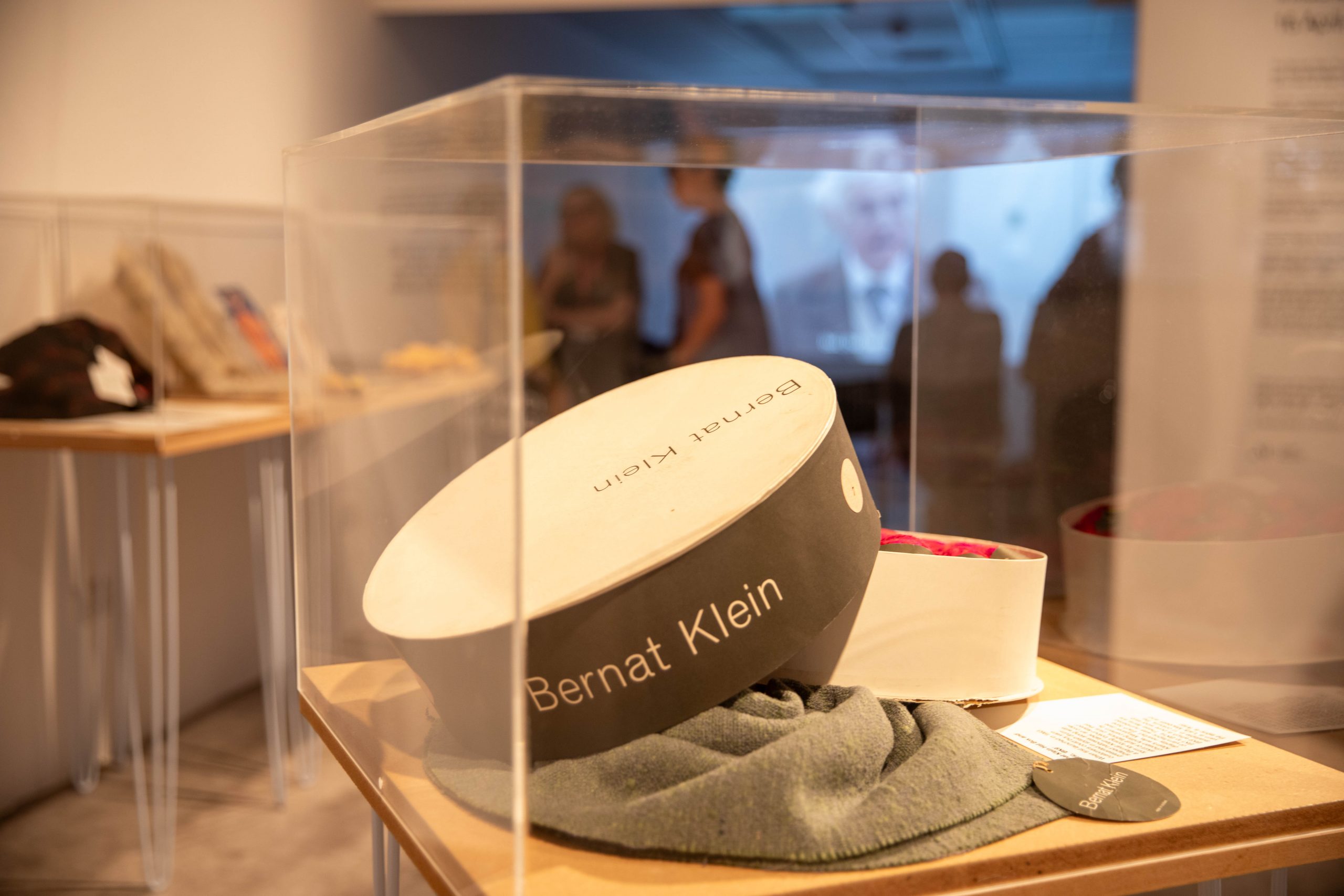 A display case contains branded packaging and textile materials presented as part of a fashion and design exhibition. Gallery visitors and a projected video can be seen in the background.