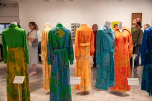 A gallery exhibition displays a series of brightly coloured patterned dresses suspended on mannequins, with visitors viewing the installation in the background. The garments feature vivid green, orange, blue and pink designs.