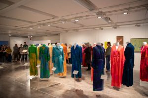 Visitors view an exhibition of suspended dresses arranged in a circular display within a gallery space. The installation presents a collection of vividly coloured fashion pieces alongside paintings and other artworks.