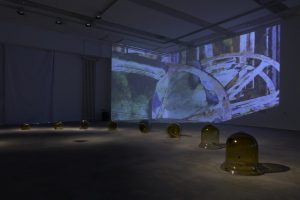 A darkened gallery installation features a row of amber glass forms across the floor, accompanied by a large abstract video projection spanning the walls. Pools of light illuminate the glass sculptures against the projected blue, green and purple imagery.