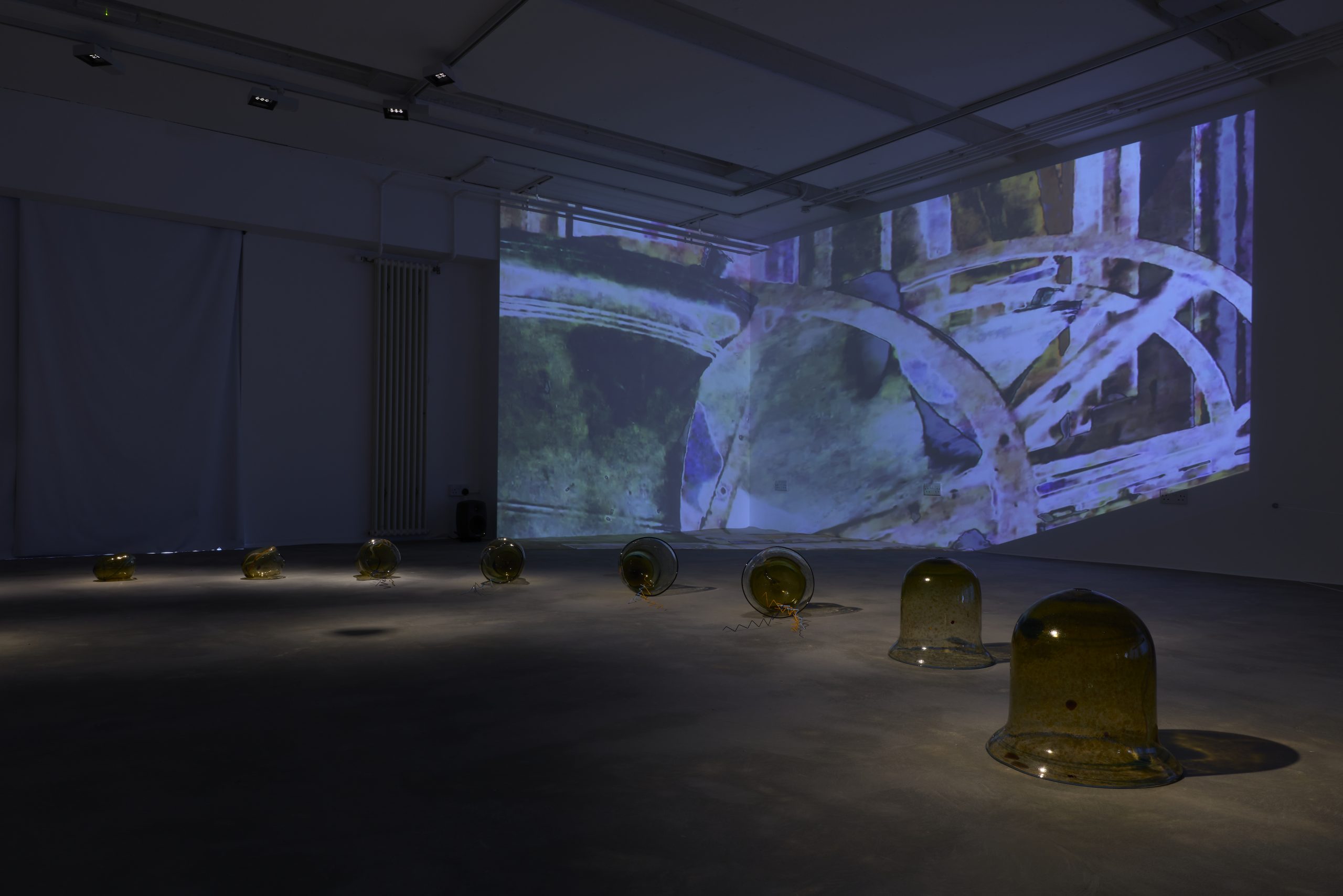 A darkened gallery installation features a row of amber glass forms across the floor, accompanied by a large abstract video projection spanning the walls. Pools of light illuminate the glass sculptures against the projected blue, green and purple imagery.