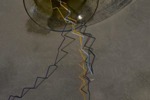 Close-up of an amber glass sculpture resting on a concrete floor, with thin angular rods in orange, blue and black extending from its opening. Strong lighting casts detailed shadows of the glass and rods across the floor.
