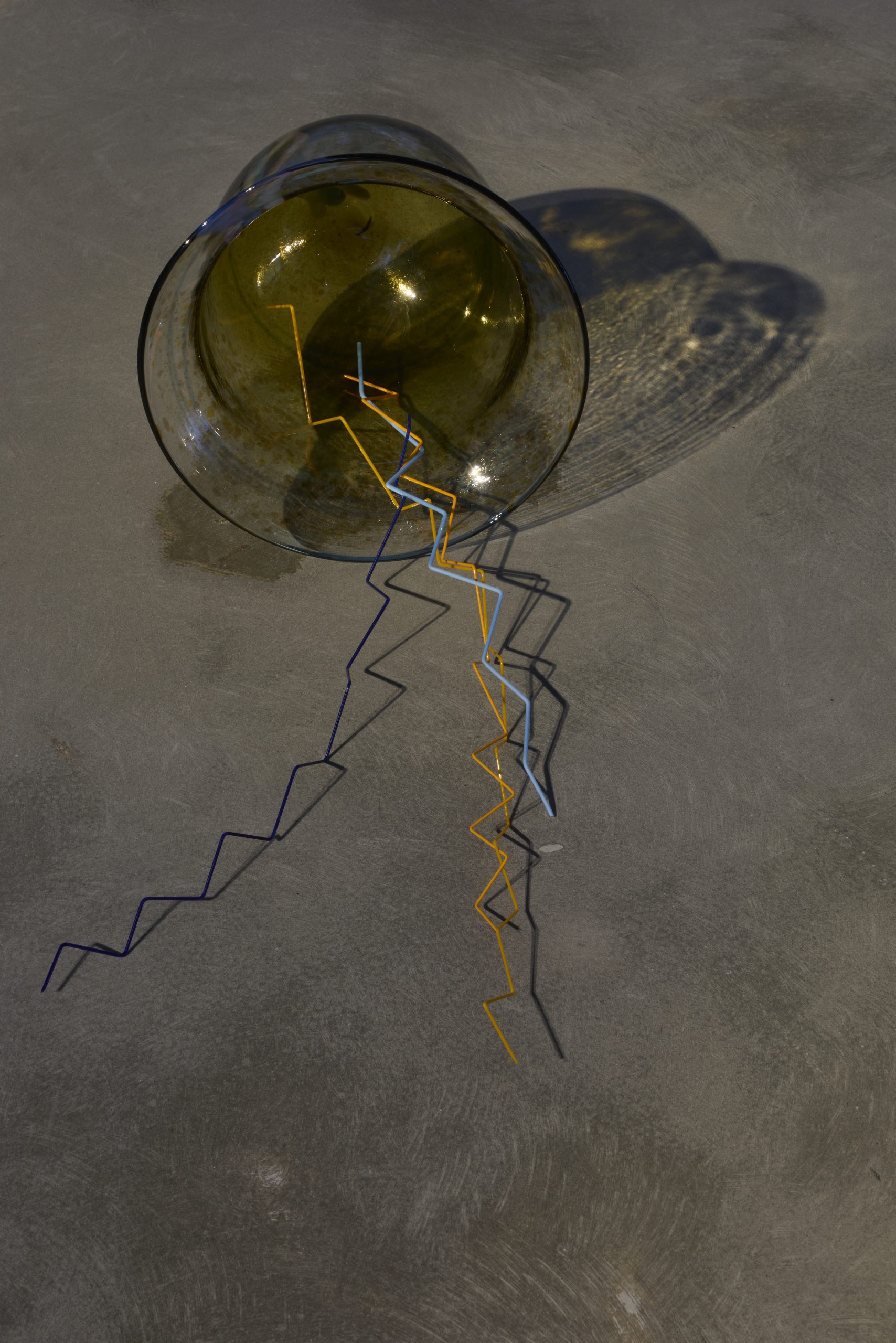 Close-up of an amber glass sculpture resting on a concrete floor, with thin angular rods in orange, blue and black extending from its opening. Strong lighting casts detailed shadows of the glass and rods across the floor.