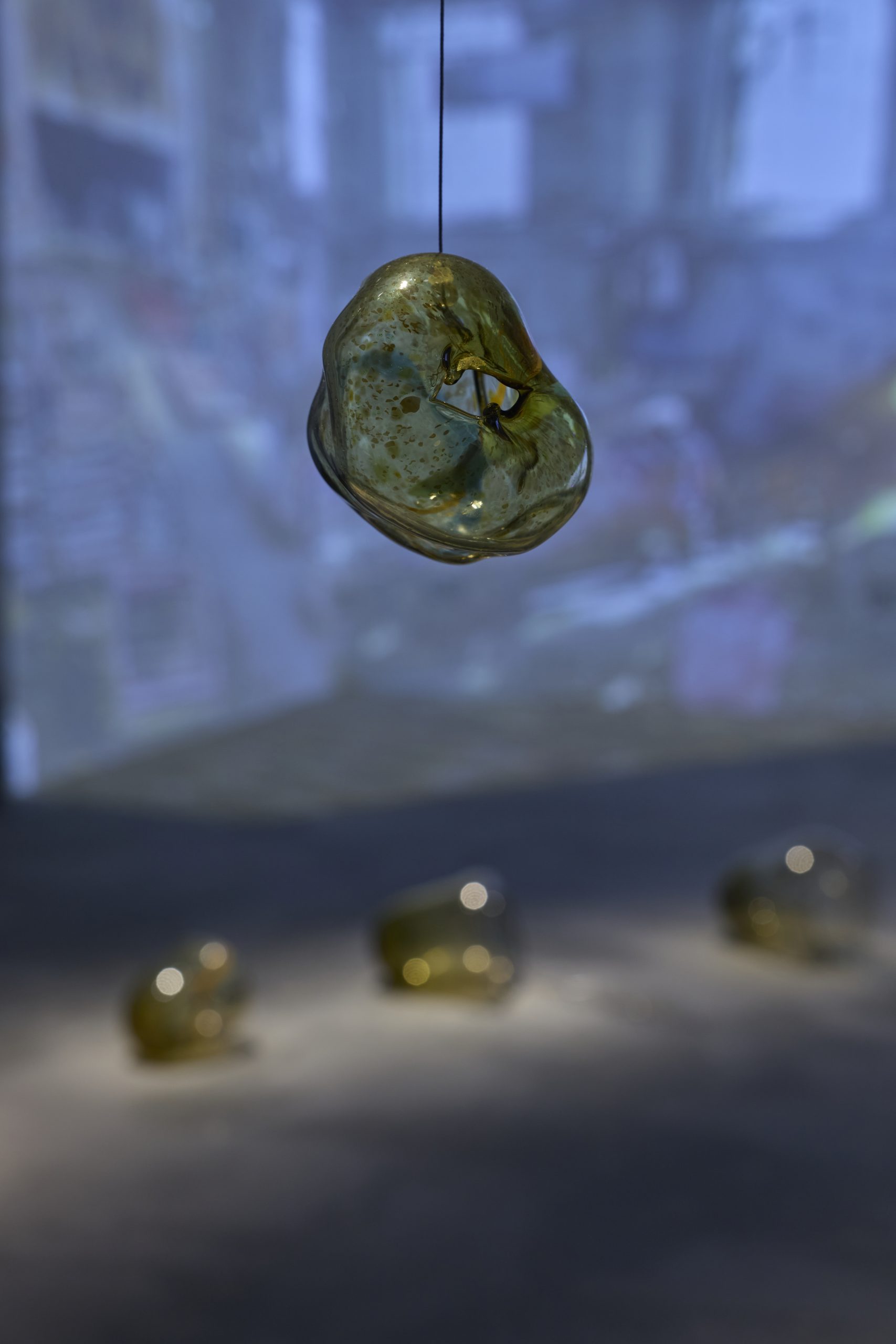 An irregular amber glass form hangs from a thin cord within a gallery installation. Additional glass forms and a softly illuminated video projection appear out of focus in the background.