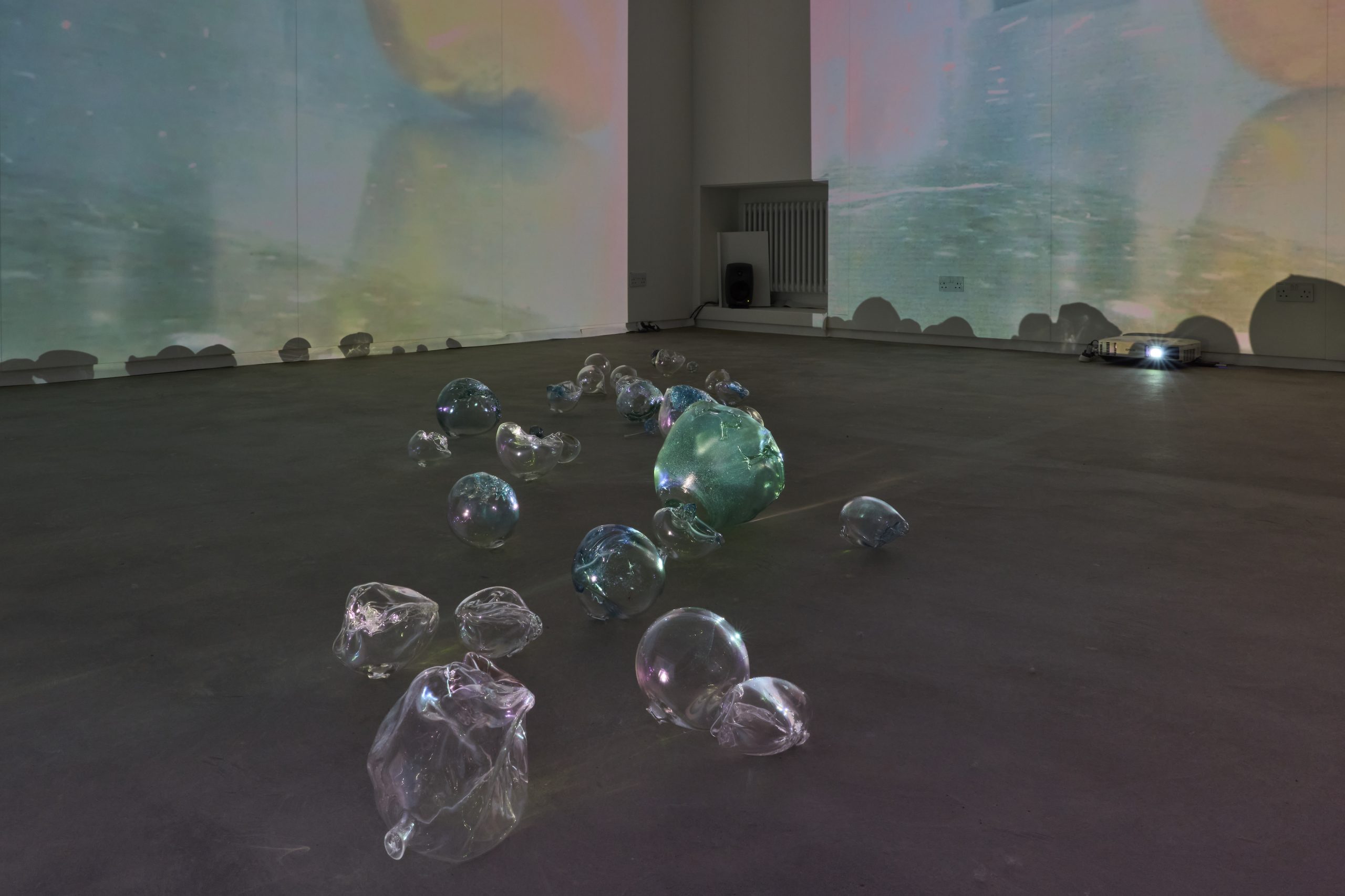 A gallery installation features numerous clear and coloured glass forms arranged across the floor beneath large pastel video projections. The glass sculptures vary in size and shape, with a large green form positioned near the centre.