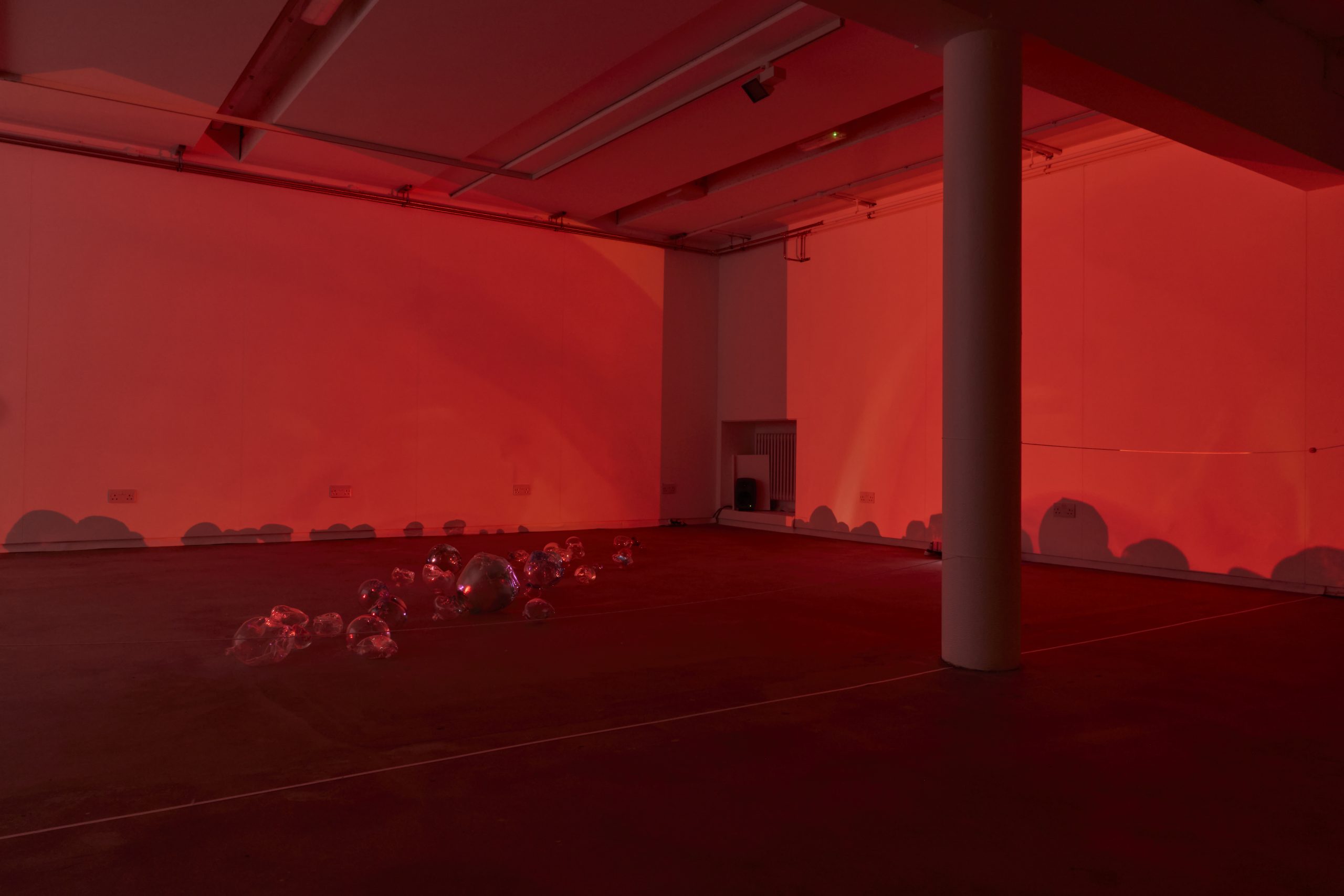 A gallery installation is illuminated entirely in deep red light, with a trail of glass forms arranged across the floor. Their shapes cast dark shadows onto the surrounding walls.