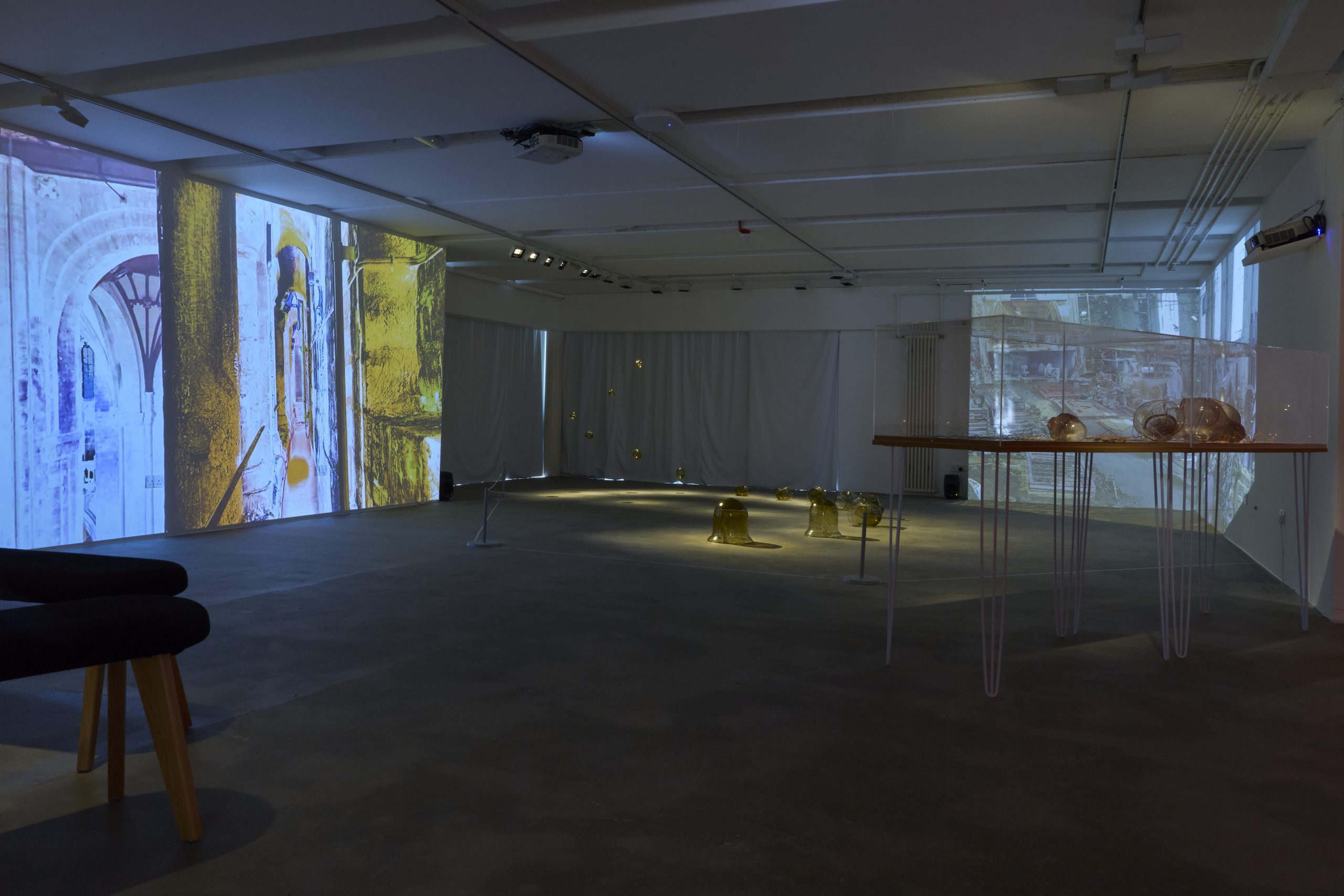 A large gallery installation combines projected architectural imagery with amber glass sculptures arranged across the floor and within a raised transparent display. Suspended glass forms and illuminated projections extend throughout the space.