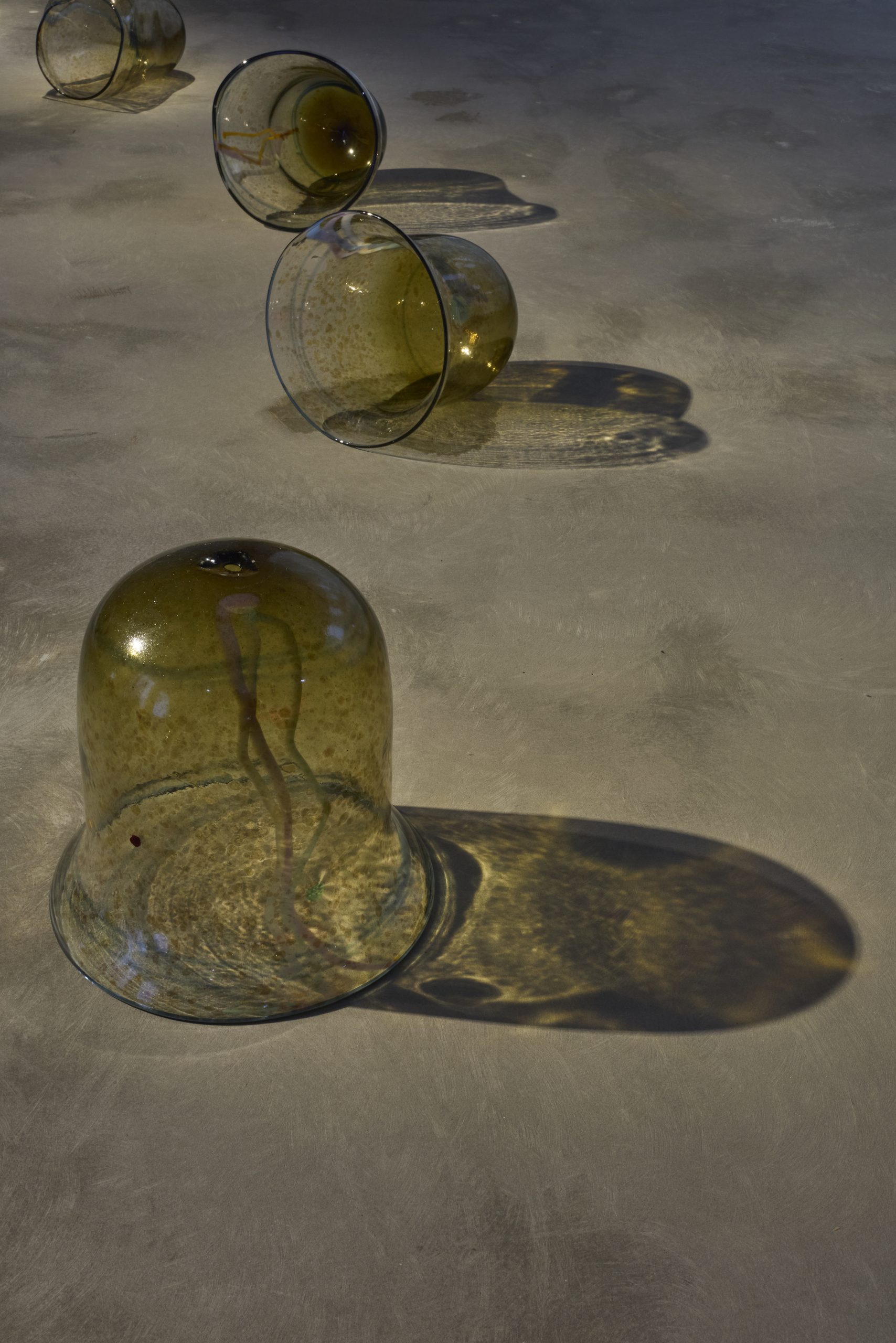 Amber glass vessels of varying shapes lie across a concrete gallery floor, casting elongated shadows under focused lighting. The closest bell-shaped vessel contains a curved coloured element within the glass.