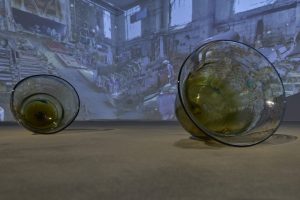 Two large amber glass vessels rest on a gallery floor in front of a projected image of an industrial interior. The translucent glass catches and reflects light from the projection.