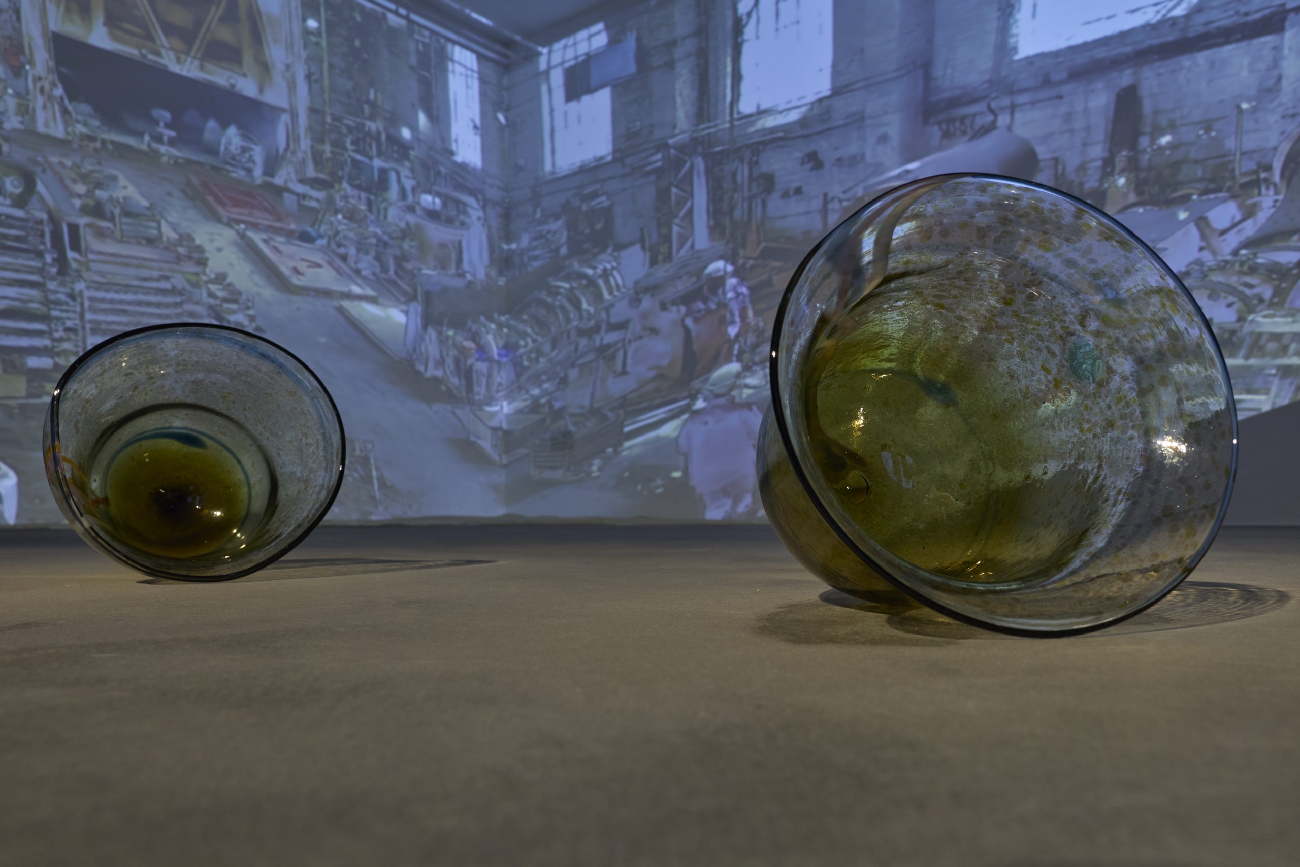 Two large amber glass vessels rest on a gallery floor in front of a projected image of an industrial interior. The translucent glass catches and reflects light from the projection.