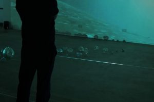 A visitor stands in silhouette within a dark gallery, viewing an immersive blue-green video projection. Small glass sculptures are arranged across the floor beneath the projected imagery.